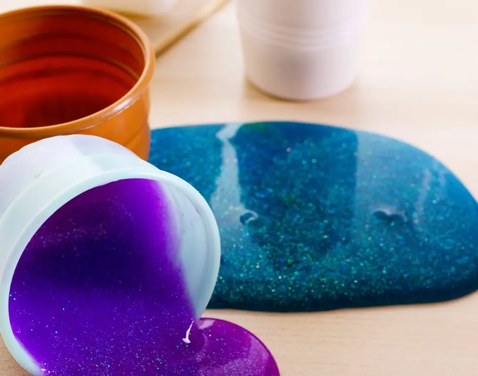 Making slime by yourself. Ingredients for making bright glittering slime at home. Violet slime spills on a table from plastic container.